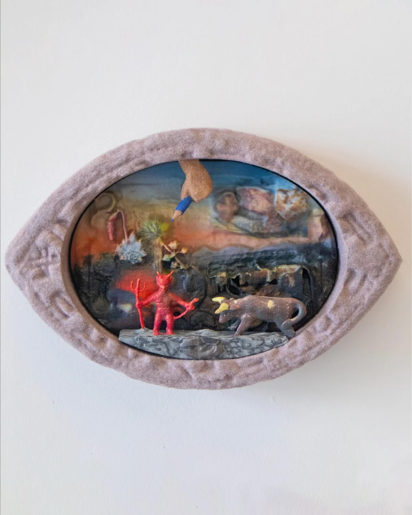 Eye-shaped wall object with a thick felt rim: inside, a small red devil figurine with a trident faces a bull on a stone ledge, beneath a hand holding a blue colored pencil descending from above